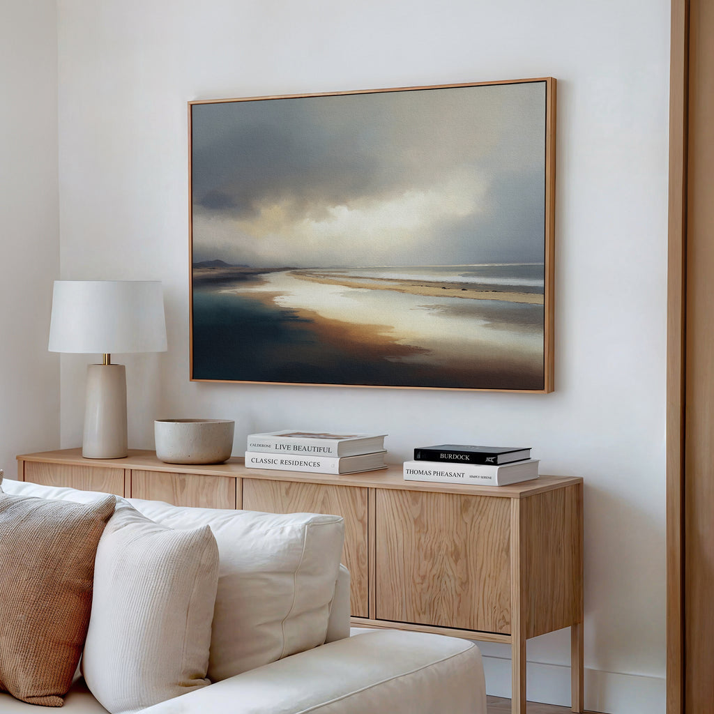 A minimalist living room features a wooden sideboard with books, a lamp, and a bowl beneath the large Tidal Reverie Canvas Art - Horizontal depicting a serene beach. A white sofa in the foreground adds to the calm, inviting atmosphere.