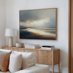 A minimalist living room features a wooden sideboard with books, a lamp, and a bowl beneath the large Tidal Reverie Canvas Art - Horizontal depicting a serene beach. A white sofa in the foreground adds to the calm, inviting atmosphere.