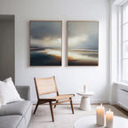 A modern, minimalist living room with a light gray sofa, wooden chair, and small white side table holding a lit candle. Above the sofa hangs Tidal Reverie - Set of 2 coastal canvas prints. Soft natural light streams through a large window.