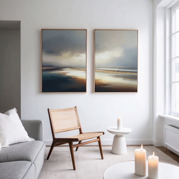 A modern, minimalist living room with a light gray sofa, wooden chair, and small white side table holding a lit candle. Above the sofa hangs Tidal Reverie - Set of 2 coastal canvas prints. Soft natural light streams through a large window.