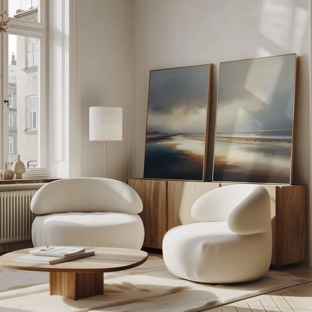 A bright, modern living room features two round white armchairs, a low wooden coffee table, and Tidal Reverie - Set of 2 canvas prints on a wood sideboard beneath tall windows with natural light.