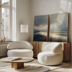 A bright, modern living room features two round white armchairs, a low wooden coffee table, and Tidal Reverie - Set of 2 canvas prints on a wood sideboard beneath tall windows with natural light.
