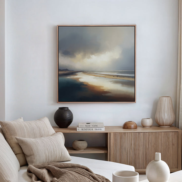 A cozy living room features a beige sofa with striped cushions, a wooden sideboard lined with books and vases, and the Tidal Reverie Canvas Art - Square—a large coastal wall art print of a serene beach scene—hanging on the wall.