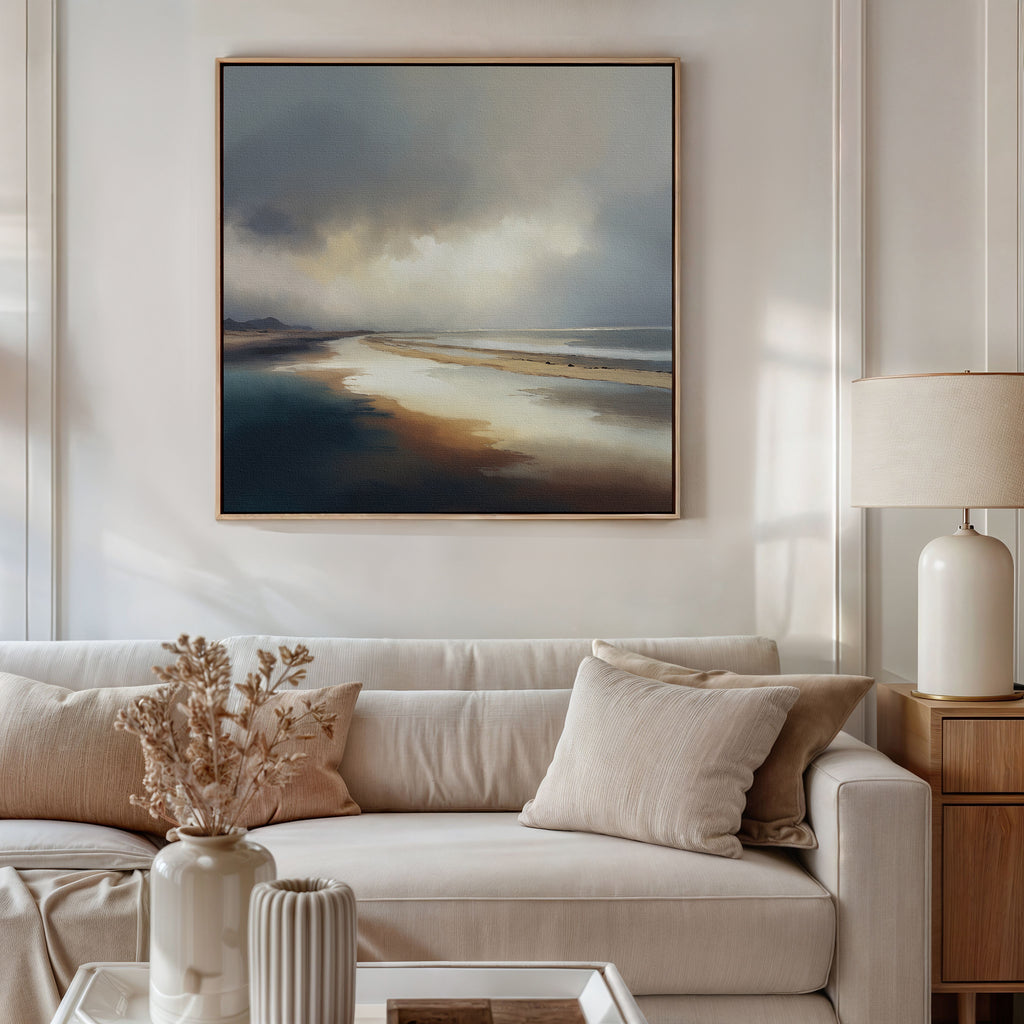 A stylish living room with a beige sofa, neutral cushions, dried flowers in a vase, and a table lamp. Above the sofa hangs the Tidal Reverie Canvas Art - Square, a large framed print showcasing a tranquil coastal scene.
