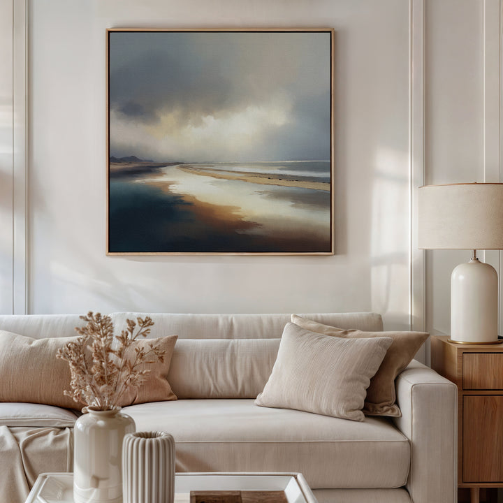 A stylish living room with a beige sofa, neutral cushions, dried flowers in a vase, and a table lamp. Above the sofa hangs the Tidal Reverie Canvas Art - Square, a large framed print showcasing a tranquil coastal scene.