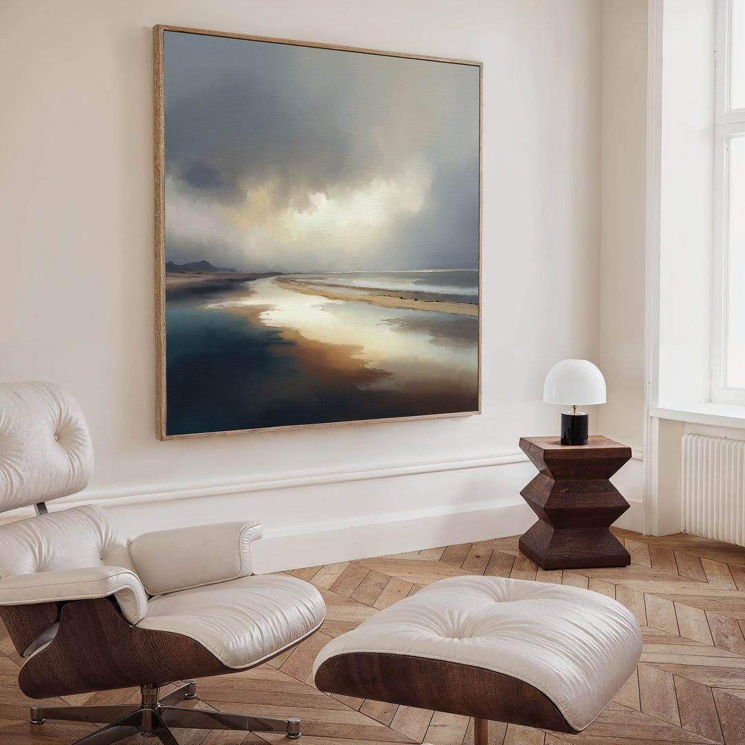 A cozy room features a white leather lounge chair and ottoman on herringbone wood flooring. Above, the Tidal Reverie Canvas Art - Square adds coastal charm, while a wooden side table with a white lamp stands by the tall window.