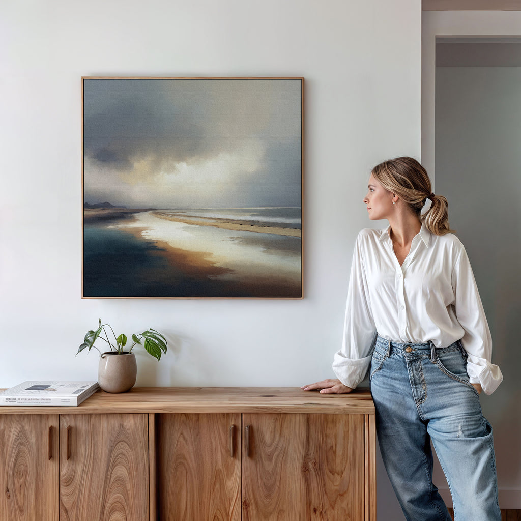 A woman in a white blouse and jeans stands beside a wooden sideboard, looking at the Tidal Reverie Canvas Art - Square, featuring a cloudy seashore scene. A plant and a book sit on the sideboard beneath the artwork.