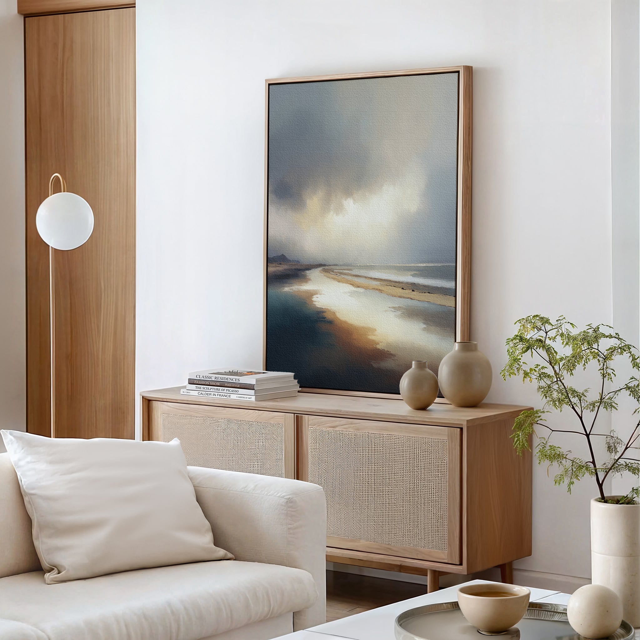 A modern living room with a light wood sideboard, neutral decor, the Tidal Reverie Canvas Art - Vertical on top, a small plant, ceramic vases, stacked books, a round wall lamp, and a white sofa with a cushion in front.