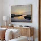 A modern living room features a light wood sideboard topped with stacked books, a white lamp, and decorative bowl. Above, the Tidal Whispers Canvas Art - Horizontal showcases a coastal marsh at sunset. A cream sofa in front completes the peaceful decor.