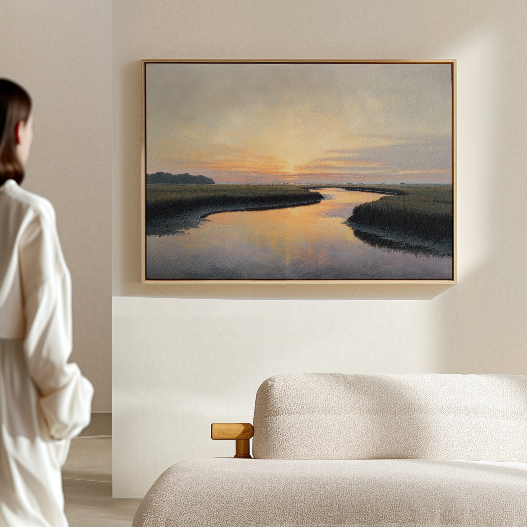 A person stands in a bright, minimal room admiring the Tidal Whispers Canvas Art - Horizontal, a serene sunset print of a coastal marsh above a beige sofa, bringing peaceful decor to the crisp white wall.