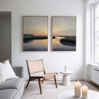 A modern living room with a wooden chair, white sofa, lit candles, and Tidal Whispers - Set of 2 large sunset-over-water canvas prints on a white wall near a tall window.