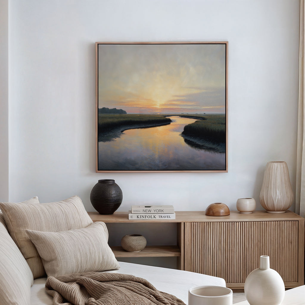 A serene living room with a light couch, neutral pillows, and a wooden sideboard. Above minimal decor and books hangs the Tidal Whispers Canvas Art - Square, depicting a coastal marsh sunset over a winding river.