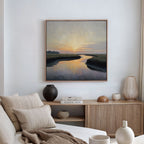 A serene living room with a light couch, neutral pillows, and a wooden sideboard. Above minimal decor and books hangs the Tidal Whispers Canvas Art - Square, depicting a coastal marsh sunset over a winding river.