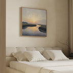 A neatly made bed with white bedding sits beneath the Tidal Whispers Canvas Art - Square, depicting a sunset over a river, hung on a beige wall in a softly lit, minimalist bedroom.