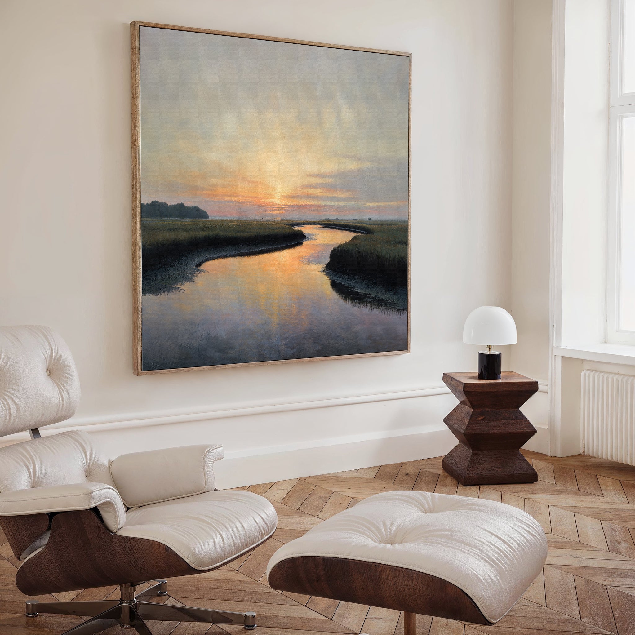 A cozy, modern living room features a white lounge chair and ottoman, a geometric wooden side table with a small white lamp, and the Tidal Whispers Canvas Art - Square as sunset-inspired wall decor above the serene space.