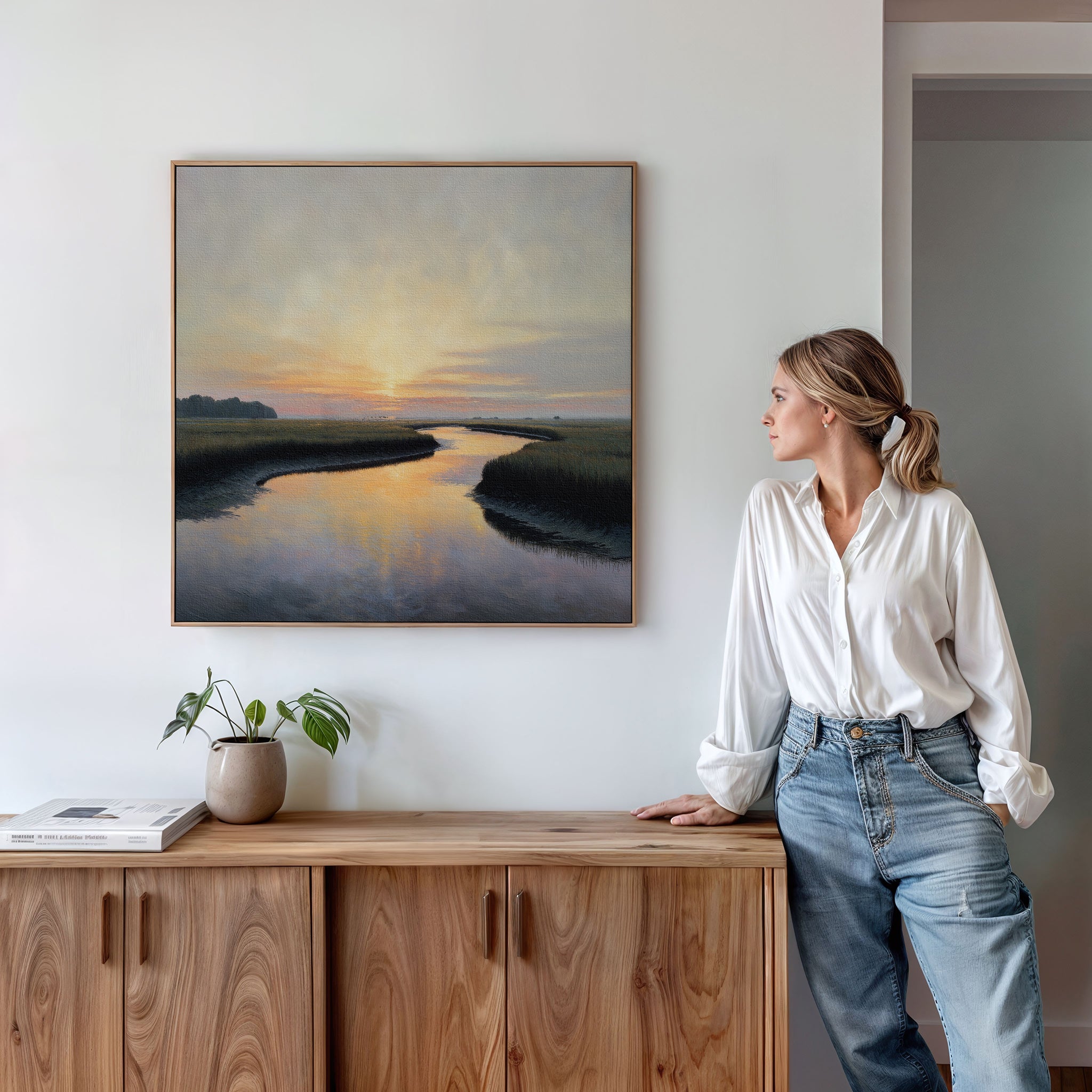 A woman in a white shirt and jeans admires the Tidal Whispers Canvas Art - Square, a framed sunset river landscape, hanging on a white wall above a wooden cabinet with a plant and books.