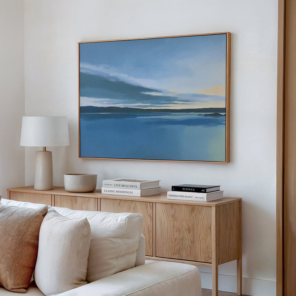 A minimalist living room features a light wood sideboard, stacked books, a table lamp, and the Tranquil Reflections Canvas Art - Horizontal above a light sofa, creating calm bedroom decor vibes with its serene lake and soft clouds scene.