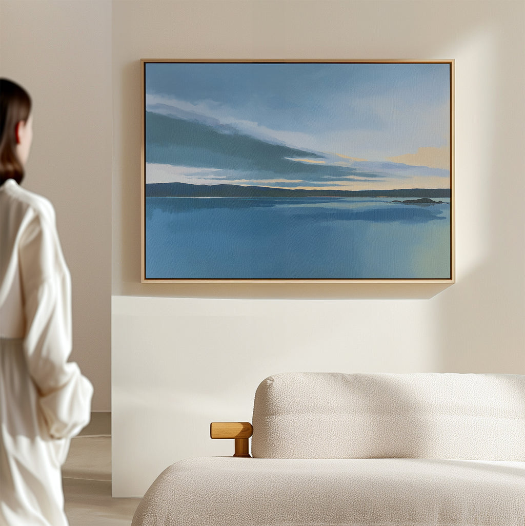 A person in a white top stands in a minimal, light-filled room, admiring the Tranquil Reflections Canvas Art - Horizontal—a blue and yellow seascape—framed above a textured beige sofa, adding calm to the bedroom decor.