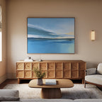 A modern living room features a wooden sideboard, coffee table with book and vase, light sofas, and the Tranquil Reflections Canvas Art - Horizontal. Soft lighting and neutral tones enhance the serene space with a wall sconce.