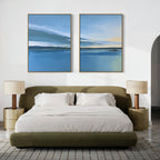 A modern bedroom with a low olive-green bed, white bedding, two gold nightstands with matching lamps, and Tranquil Reflections - Set of 2 artwork above the bed. A gray patterned rug covers the floor.