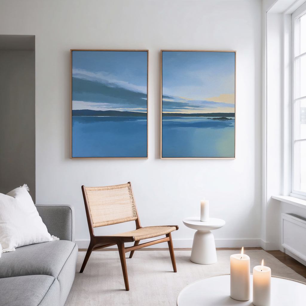 A modern, minimalist living room features a gray sofa, wooden chair, round white side table, and lit candles. Above the chair hang two large "Tranquil Reflections - Set of 2" artwork pieces with blue and yellow tones.