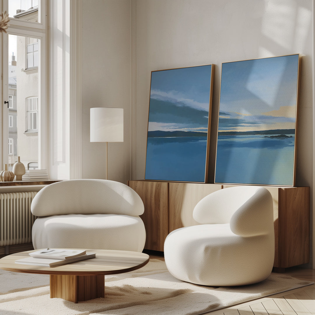 A modern living room with two white, curved armchairs, a low wooden coffee table, and the "Tranquil Reflections - Set of 2" wall art above a wooden sideboard near large windows that flood the space with natural light.