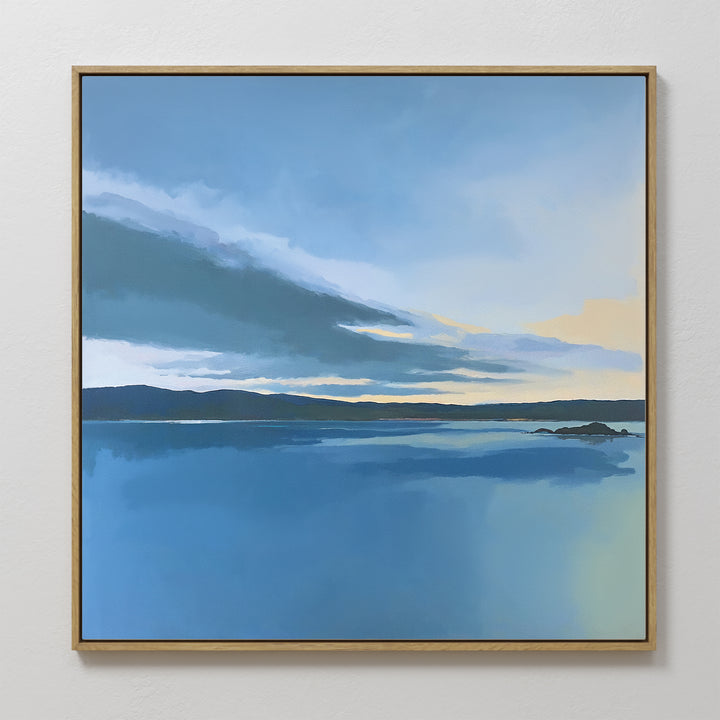 The Tranquil Reflections Canvas Art - Square features a calm blue lake with a distant shoreline beneath a blue, gray, and yellow sky, evoking sunrise or sunset. Serene and softly rendered, it’s ideal for tranquil bedroom decor.