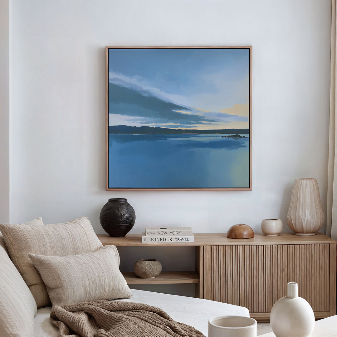 A serene living room with a beige sofa, neutral cushions, and a wooden sideboard decorated with vases and books. Above, the Tranquil Reflections Canvas Art - Square adds calming beauty to the white wall as peaceful decor.