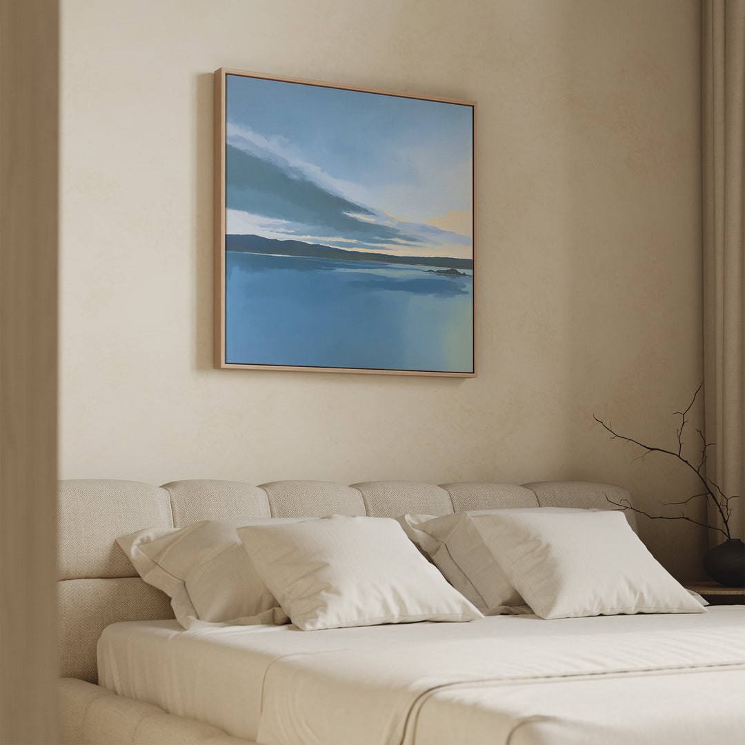 A minimal bedroom with a beige upholstered bed, white bedding, and pillows. The Tranquil Reflections Canvas Art - Square in blue and yellow hangs above the bed, enhancing the serene decor alongside a bare branch on the side table.