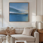 A modern living room with a beige sofa, matching cushions, a side table with a lamp, dried flowers in a vase, and the Tranquil Reflections Canvas Art - Square—blue-toned landscape wall art—hanging above.