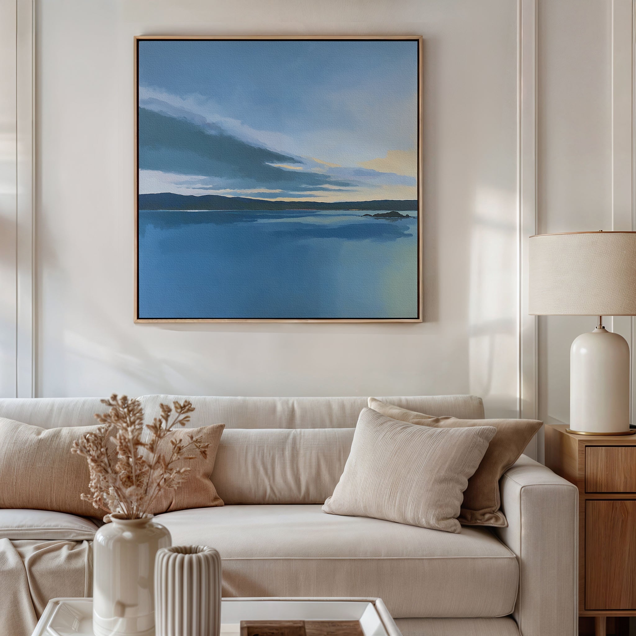 A modern living room with a beige sofa, matching cushions, a side table with a lamp, dried flowers in a vase, and the Tranquil Reflections Canvas Art - Square—blue-toned landscape wall art—hanging above.