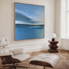 A modern, minimalist living room with a white lounge chair and ottoman, a small wooden side table with a white lamp, and serene wall art—a large Tranquil Reflections Canvas Art - Square evoking peaceful blue lakescapes.