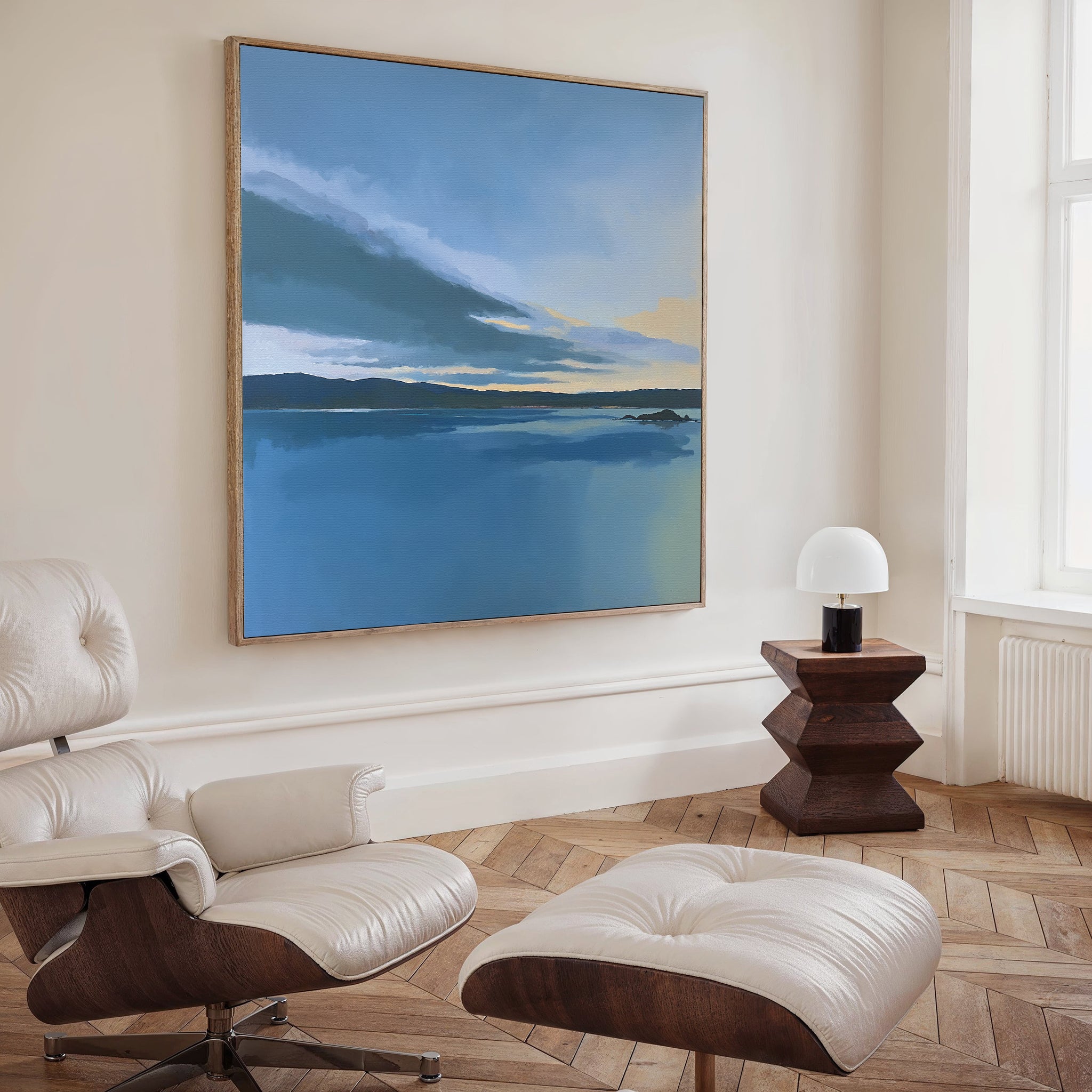 A modern, minimalist living room with a white lounge chair and ottoman, a small wooden side table with a white lamp, and serene wall art—a large Tranquil Reflections Canvas Art - Square evoking peaceful blue lakescapes.