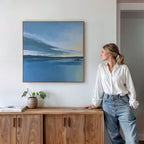 A woman in a white blouse and jeans leans against a wooden cabinet, admiring the Tranquil Reflections Canvas Art - Square, a serene landscape of lake and sky. A potted plant and books rest on the cabinet, enhancing this peaceful bedroom decor.