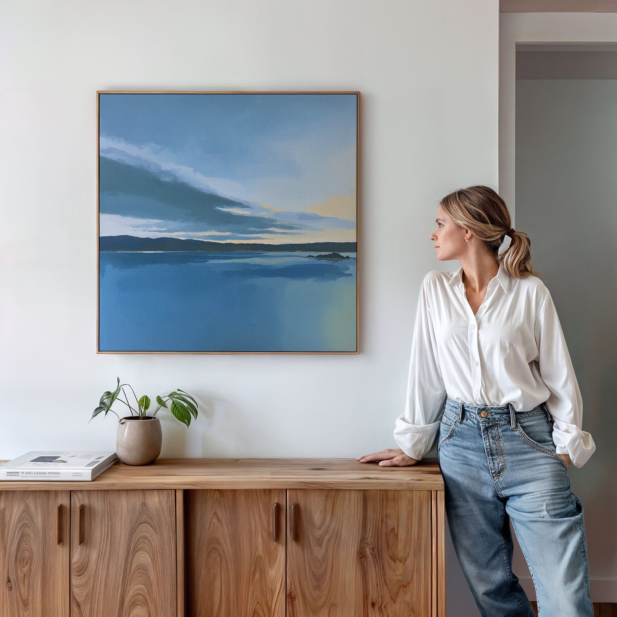 A woman in a white blouse and jeans leans against a wooden cabinet, admiring the Tranquil Reflections Canvas Art - Square, a serene landscape of lake and sky. A potted plant and books rest on the cabinet, enhancing this peaceful bedroom decor.
