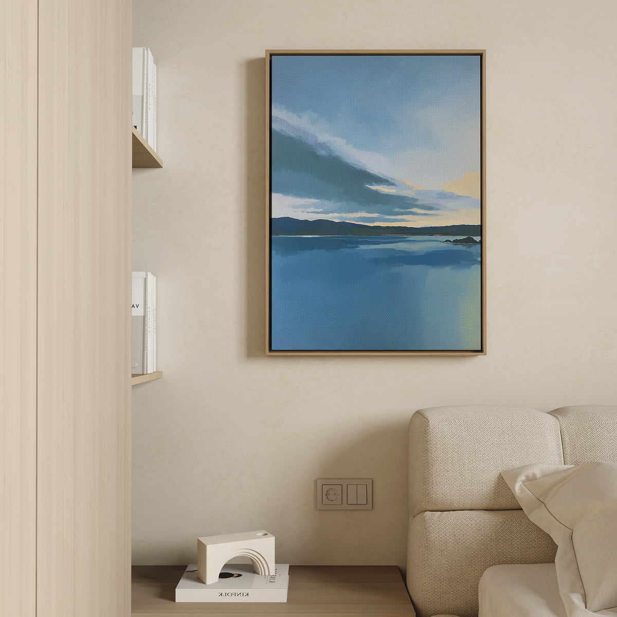 A modern living room corner with a beige sofa, light wood shelves of books, a small white geometric decor piece on the floor, and “Tranquil Reflections Canvas Art - Vertical”—a serene blue abstract painting—framed on the wall.