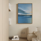 A modern living room corner with a beige sofa, light wood shelves of books, a small white geometric decor piece on the floor, and “Tranquil Reflections Canvas Art - Vertical”—a serene blue abstract painting—framed on the wall.