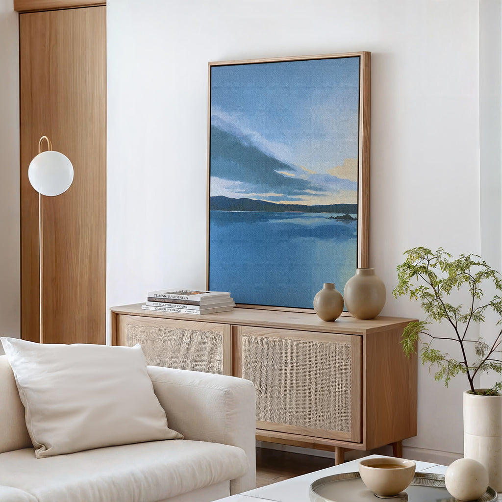 A minimalist living room with a white sofa, light wood sideboard, ceramic vases, and the Tranquil Reflections Canvas Art - Vertical featuring serene blue skies and water. A small green plant and books accent the modern, peaceful decor.