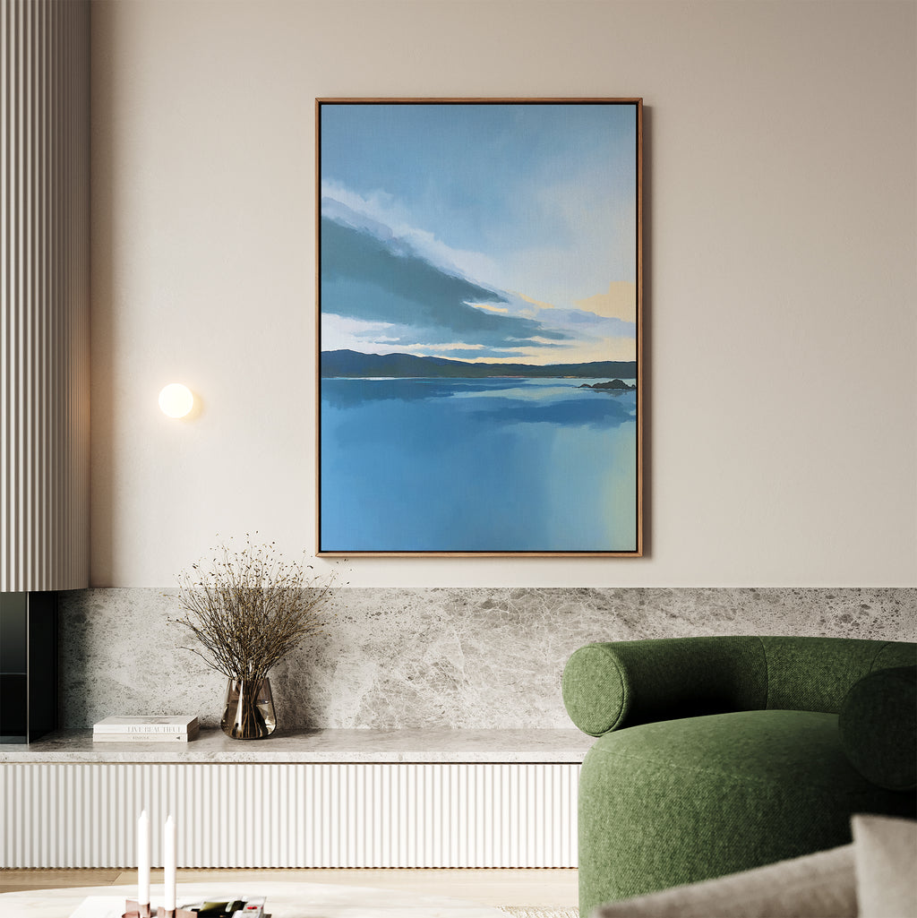 A modern living room features a green sofa, marble accents, and Tranquil Reflections Canvas Art - Vertical—a large framed blue landscape with mountains and a cloudy sky—hung on a light wall. Dried flowers sit on the shelf below.