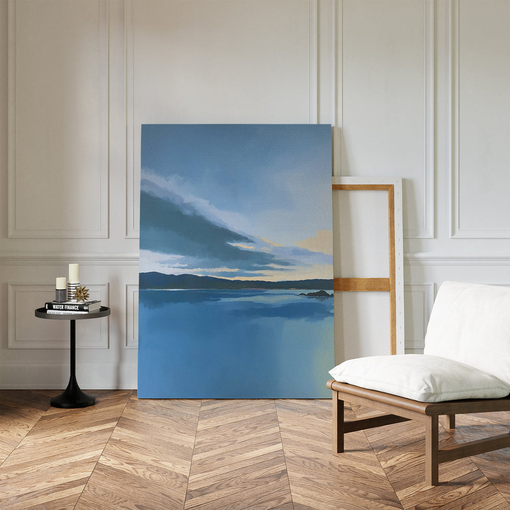 The Tranquil Reflections Canvas Art - Vertical, featuring a calm lake and pastel sky, leans against a white paneled wall next to an empty wooden frame, with a cushioned chair and candlelit table on a chevron wood floor.