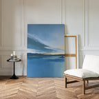The Tranquil Reflections Canvas Art - Vertical, featuring a calm lake and pastel sky, leans against a white paneled wall next to an empty wooden frame, with a cushioned chair and candlelit table on a chevron wood floor.