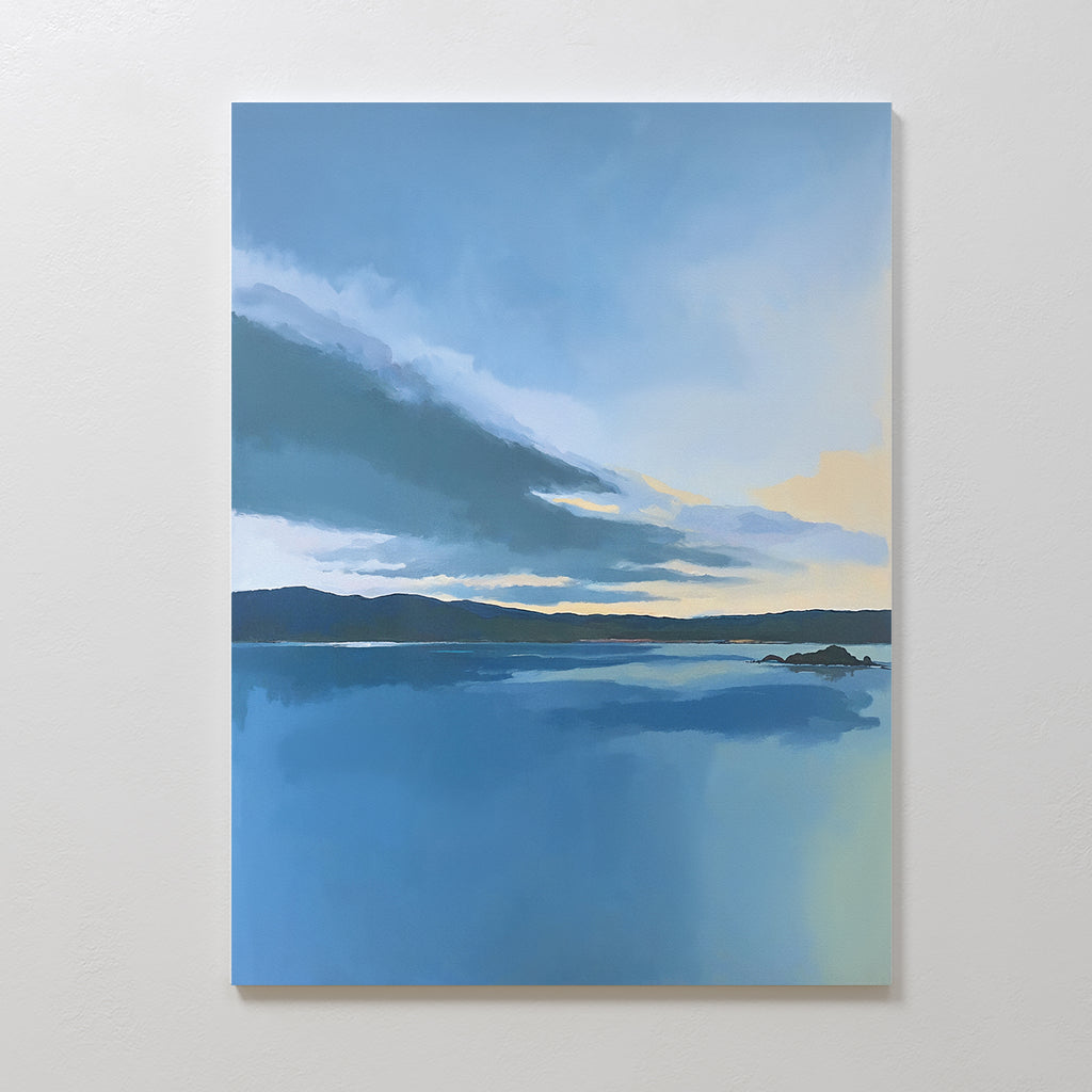 The Tranquil Reflections Canvas Art - Vertical features a serene blue lake mirroring the sky with distant hills and clouds, displayed on a plain white wall.