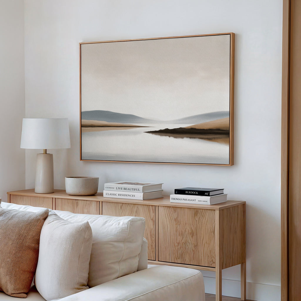A modern, minimalist living room features a wooden sideboard with stacked books, a white lamp, and a bowl. Above hangs the Tranquil Waters Canvas Art - Horizontal, displaying a misty lakeside scene in neutral tones. A white couch is partially visible.