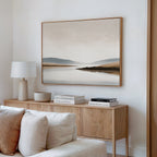 A modern, minimalist living room features a wooden sideboard with stacked books, a white lamp, and a bowl. Above hangs the Tranquil Waters Canvas Art - Horizontal, displaying a misty lakeside scene in neutral tones. A white couch is partially visible.