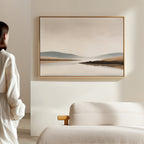 A person in a white shirt stands near a minimalist bedroom with a textured bed and wooden armrest, gazing at the Tranquil Waters Canvas Art - Horizontal, featuring a serene lakeside landscape, on the wall.