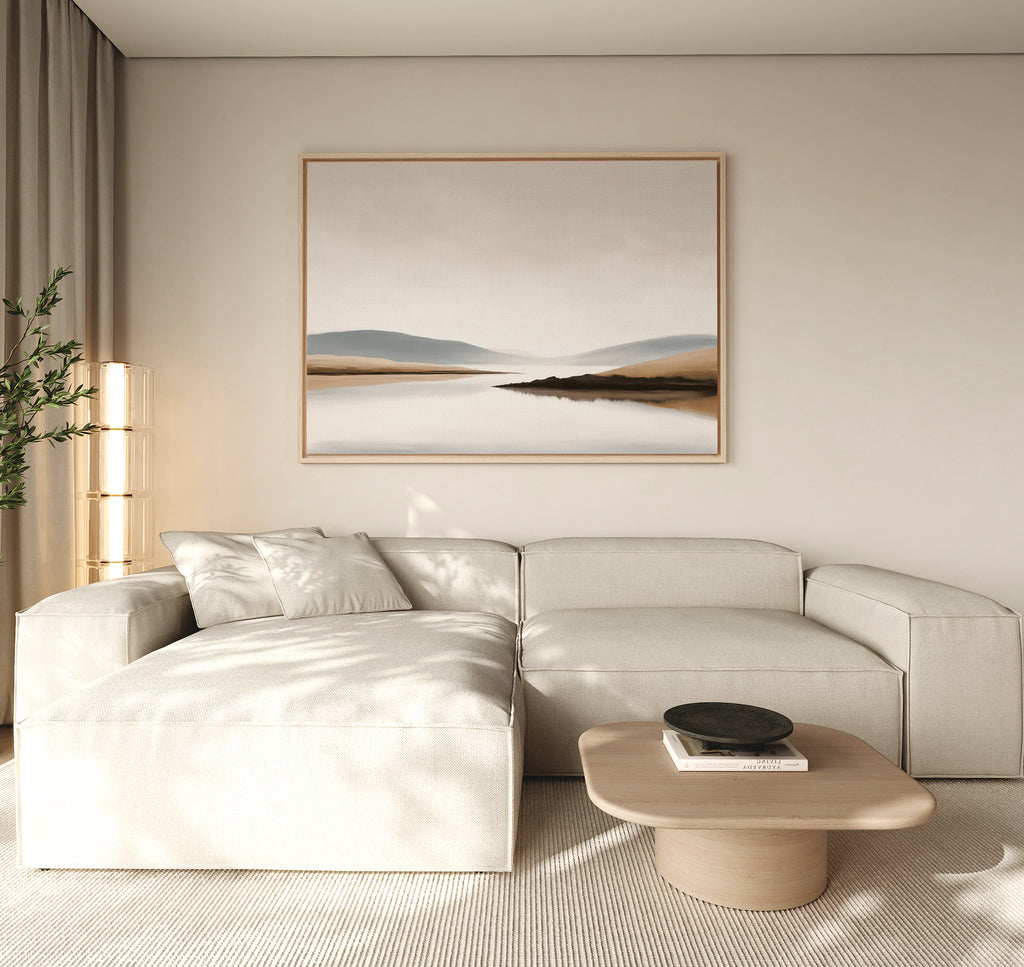 A modern living room features a light beige sectional sofa, low wooden coffee table, black decorative bowl, tall plant, and the Tranquil Waters Canvas Art - Horizontal depicting a misty lakeside landscape on the wall.