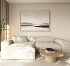 A modern living room features a light beige sectional sofa, low wooden coffee table, black decorative bowl, tall plant, and the Tranquil Waters Canvas Art - Horizontal depicting a misty lakeside landscape on the wall.