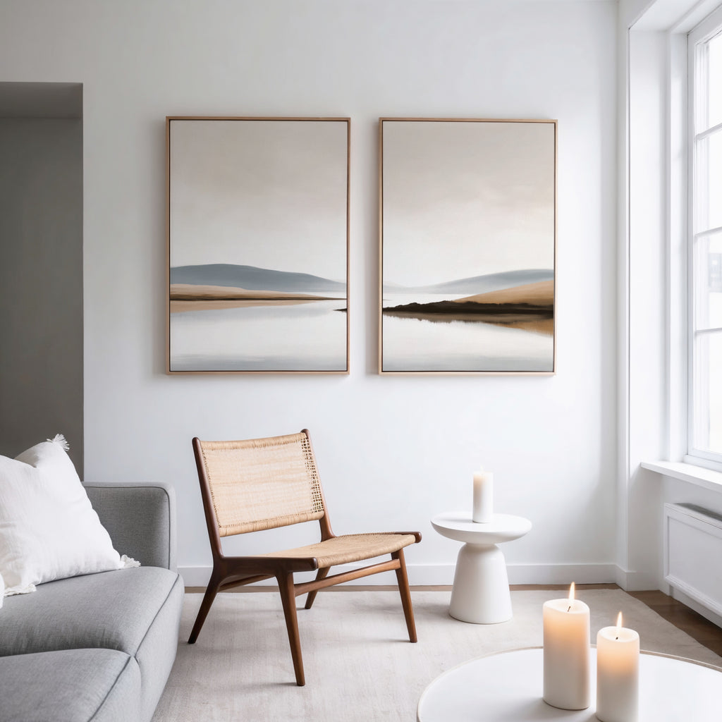 A minimalist living room with a gray sofa, wooden chair, and small round table holding a lit candle. Above hangs Tranquil Waters - Set of 2 canvas prints. Large windows fill the space with light; more candles sit on a low table.