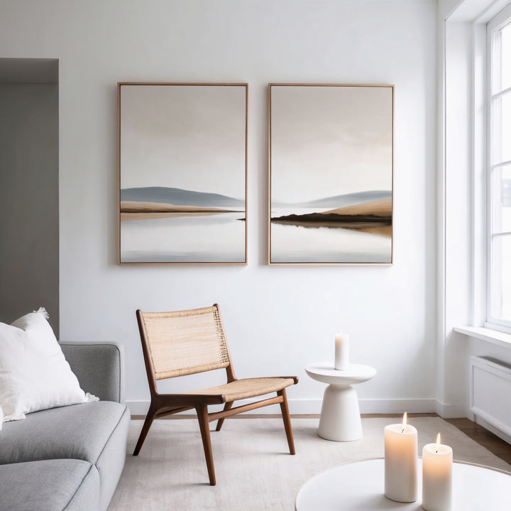 A minimalist living room with a gray sofa, wooden chair, and small round table holding a lit candle. Above hangs Tranquil Waters - Set of 2 canvas prints. Large windows fill the space with light; more candles sit on a low table.