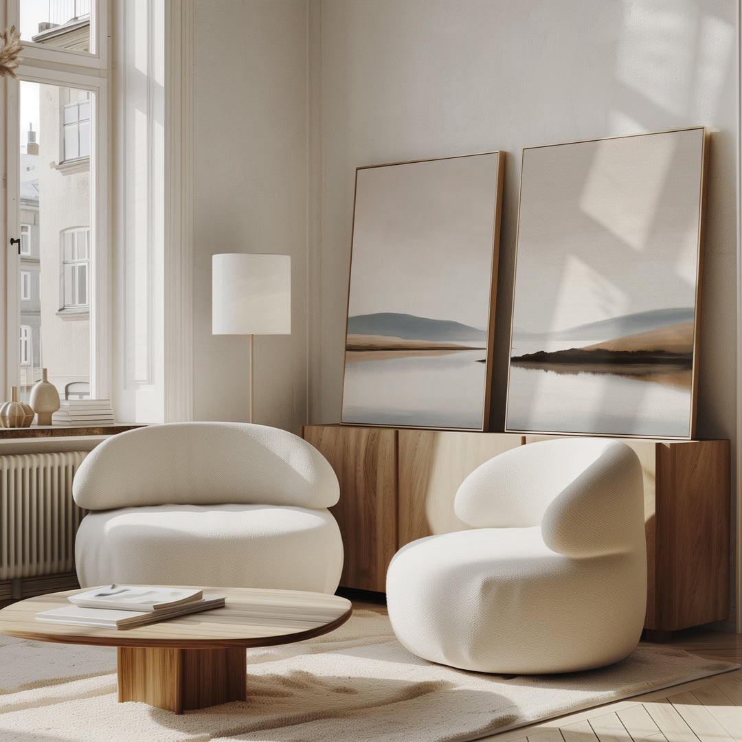 A modern, minimal living room with two rounded white armchairs, a low wood coffee table, and the Tranquil Waters - Set of 2 misty lakeside canvas prints on a wooden cabinet. Sunlight from large windows creates a calm, airy vibe.